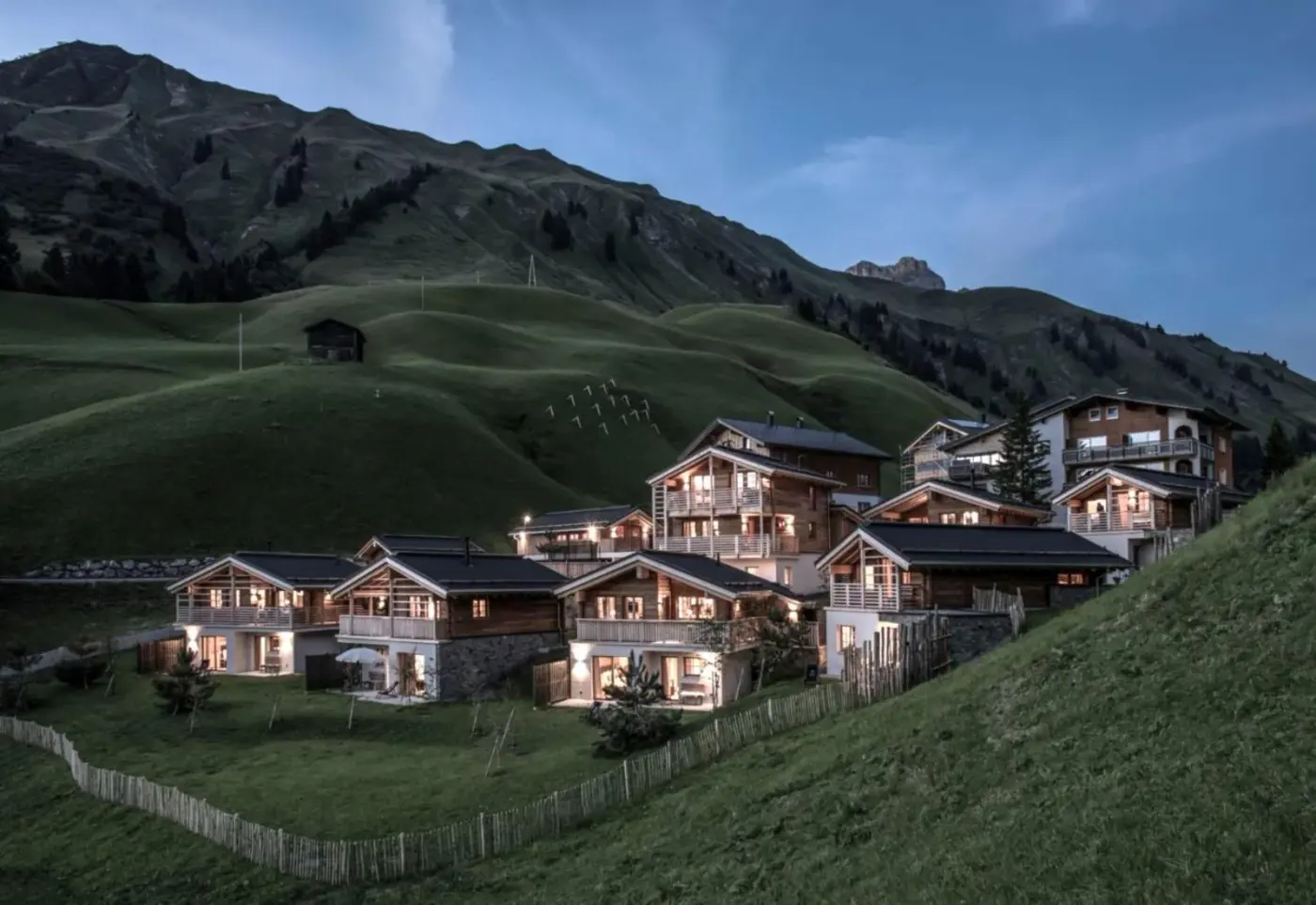 Timber chalet village illuminated at dusk on a green alpine hillside at Aadla Walser Chalets in Schröcken, Austria.