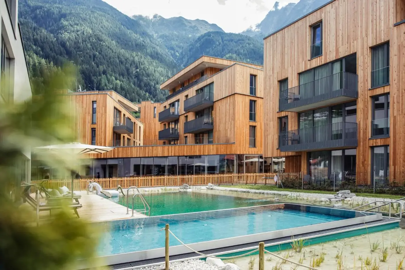 Timber resort buildings beside an outdoor swimming pool beneath alpine mountains at All-Suite Resort Ötztal in Oetz, Austria.
