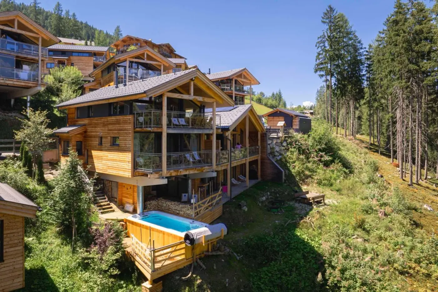 Timber chalet with a balcony and outdoor spa tub on a wooded hillside at Alpenchalets Reiteralm by ALPS RESORTS in Pichl bei Schladming, Austria.