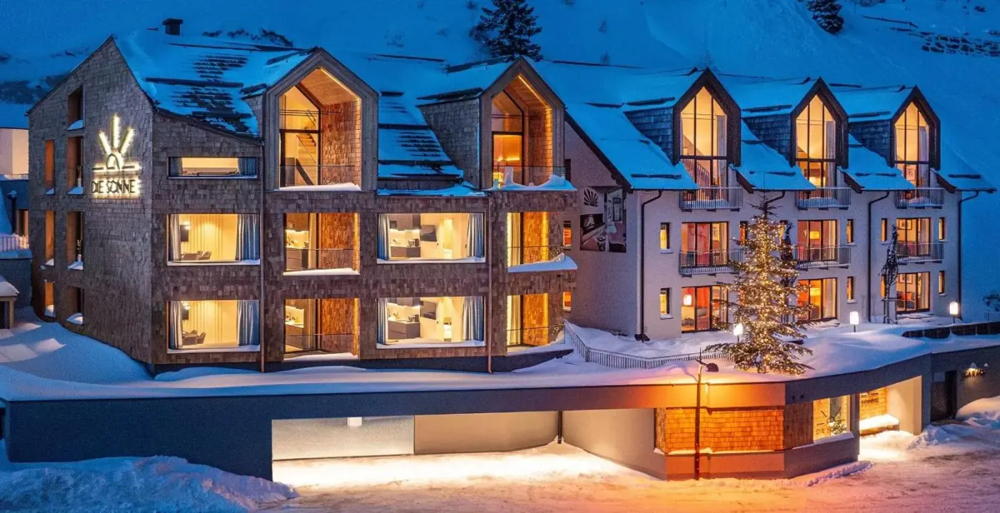 Illuminated alpine aparthotel exterior surrounded by snow at dusk at Aparthotel DIE SONNE in Obertauern, Austria.