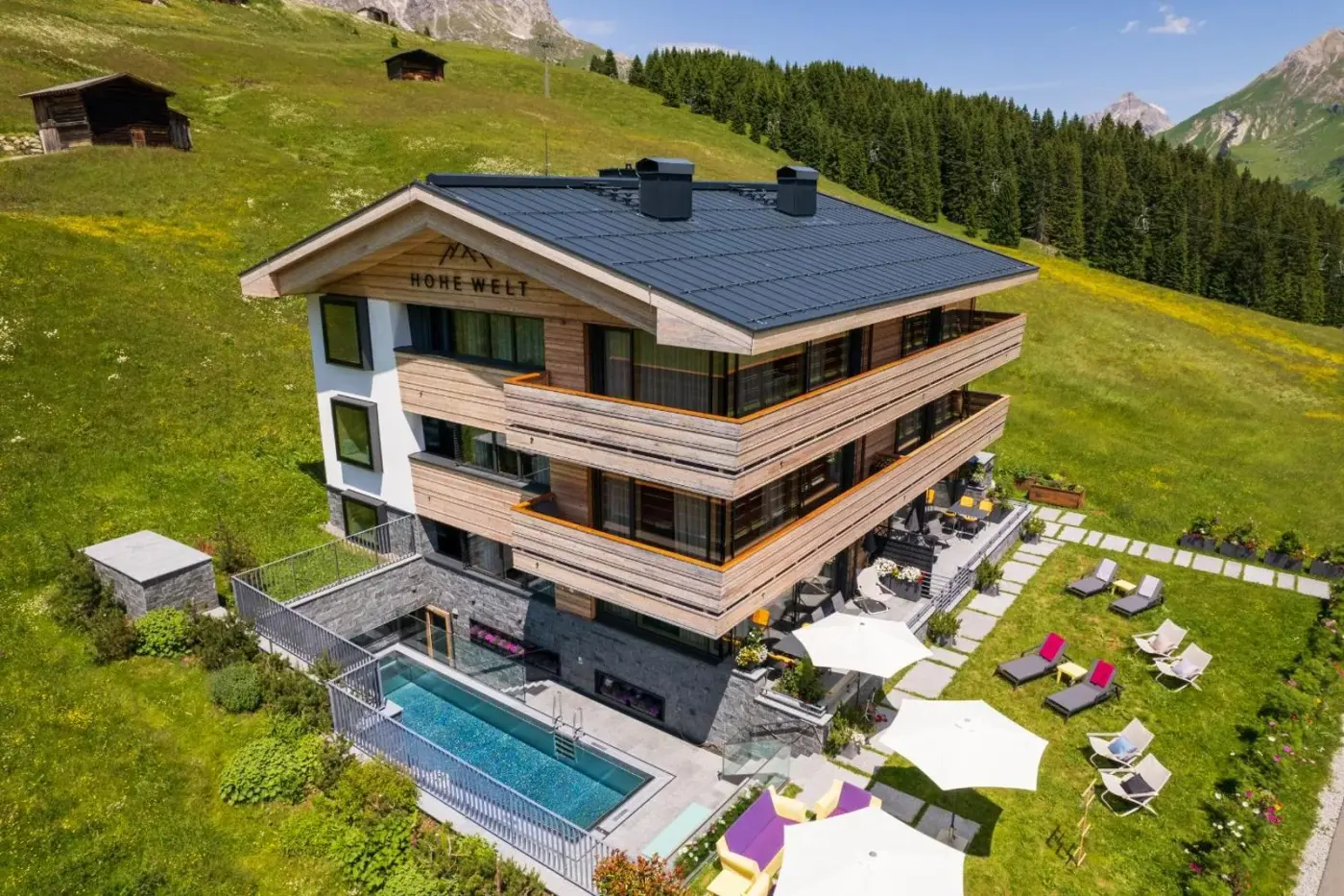 Aerial view of the timber chalet, outdoor pool and mountain meadow at Chalet Hohe Welt in Lech am Arlberg, Austria.
