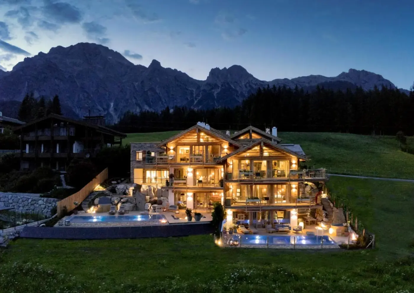 Illuminated chalet and pool terraces beneath a dark mountain skyline at GipfelKreuzLiebe SENHOOG in Leogang, Austria.