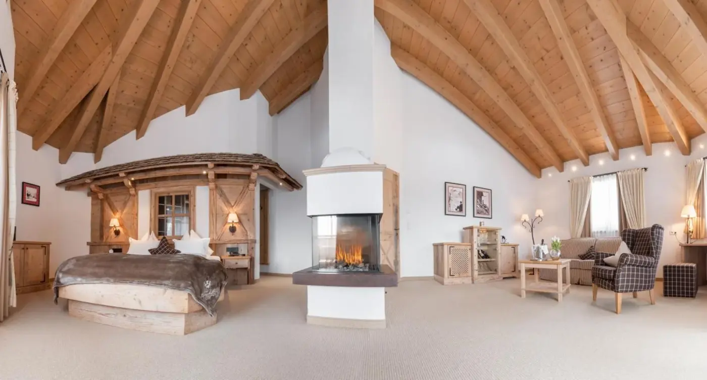 Spacious suite with a double bed and fireplace beneath a vaulted timber roof at Hotel Leitenhof in Scheffau am Wilden Kaiser, Austria.