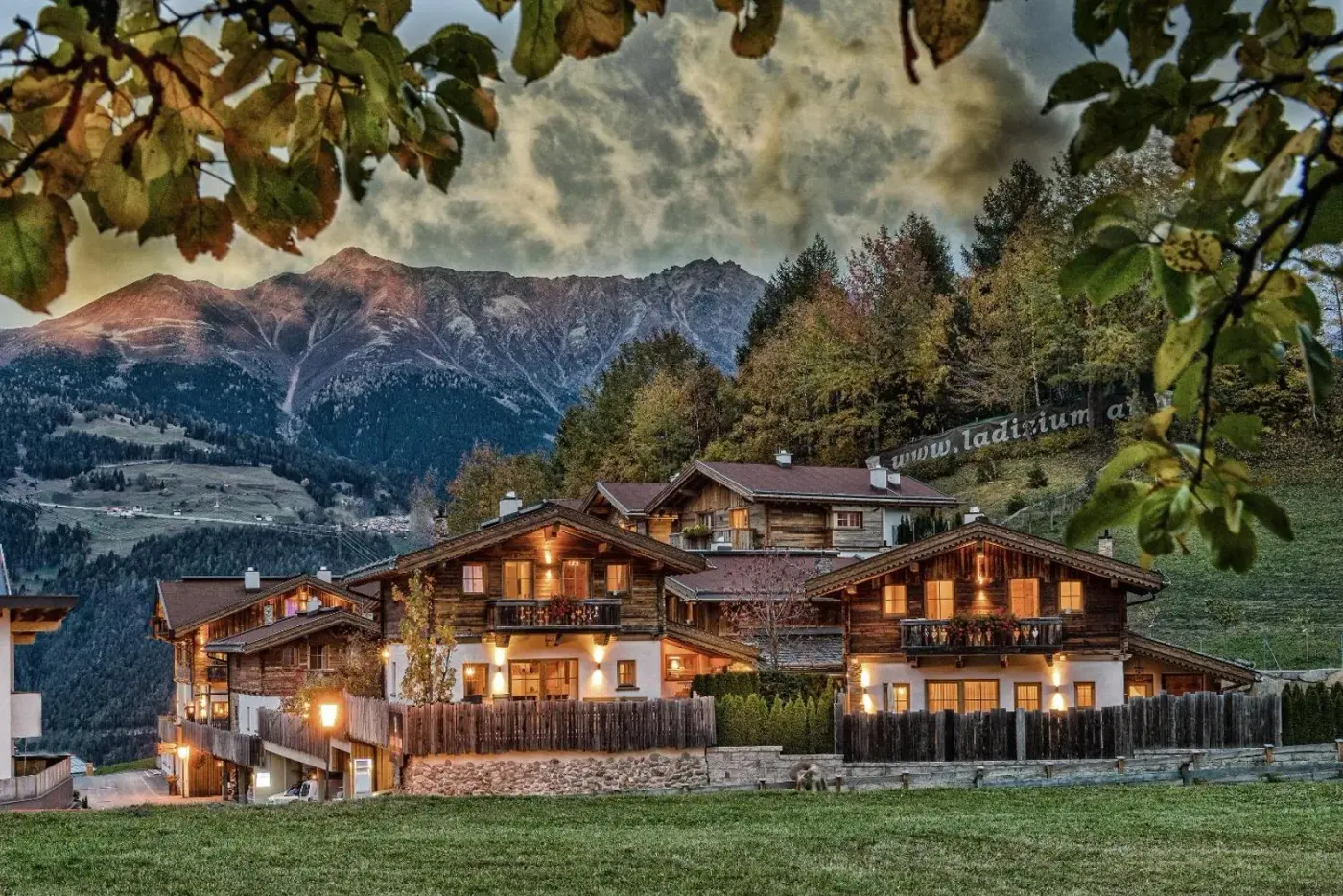 Illuminated timber chalet village beneath mountains at sunset at Hüttendorf Ladizium in Ladis, Austria.