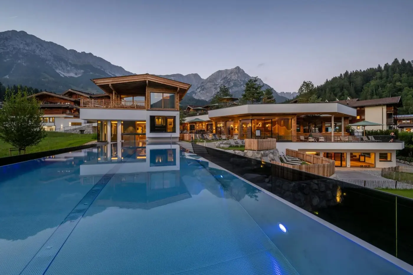 Rooftop pool reflecting timber buildings with mountains behind at dusk at Kaiserlodge in Scheffau am Wilden Kaiser, Austria.