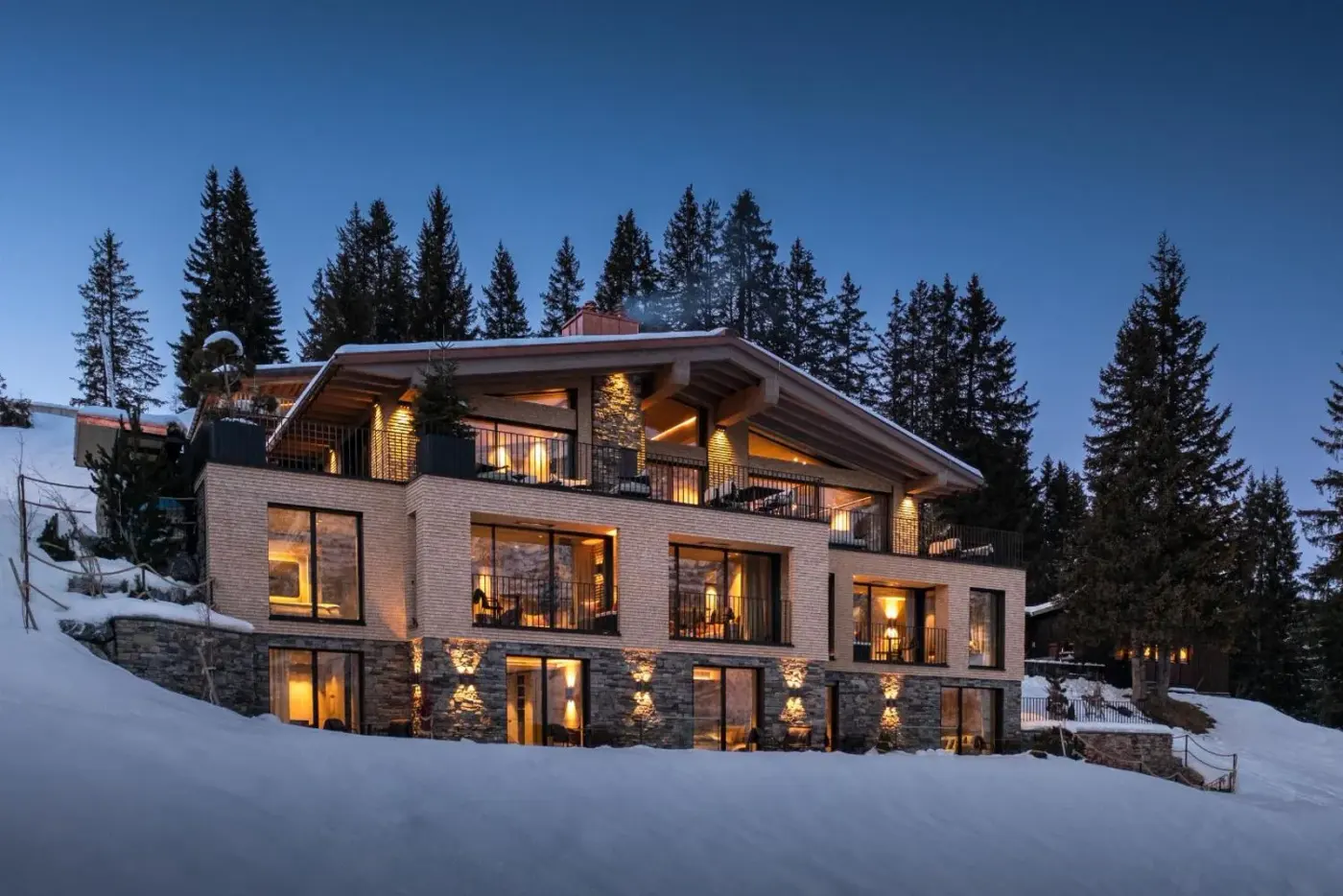 Illuminated stone and timber chalet surrounded by snow at dusk at LE CHALET Luxury Hideaway in Lech am Arlberg, Austria.