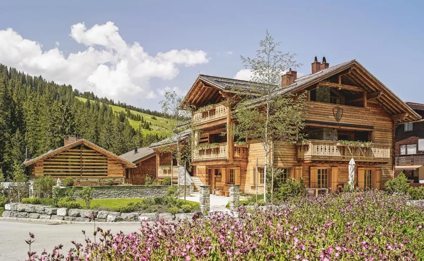 Timber chalet exterior surrounded by flowers and green alpine slopes at Lech Lodge in Lech am Arlberg, Austria.