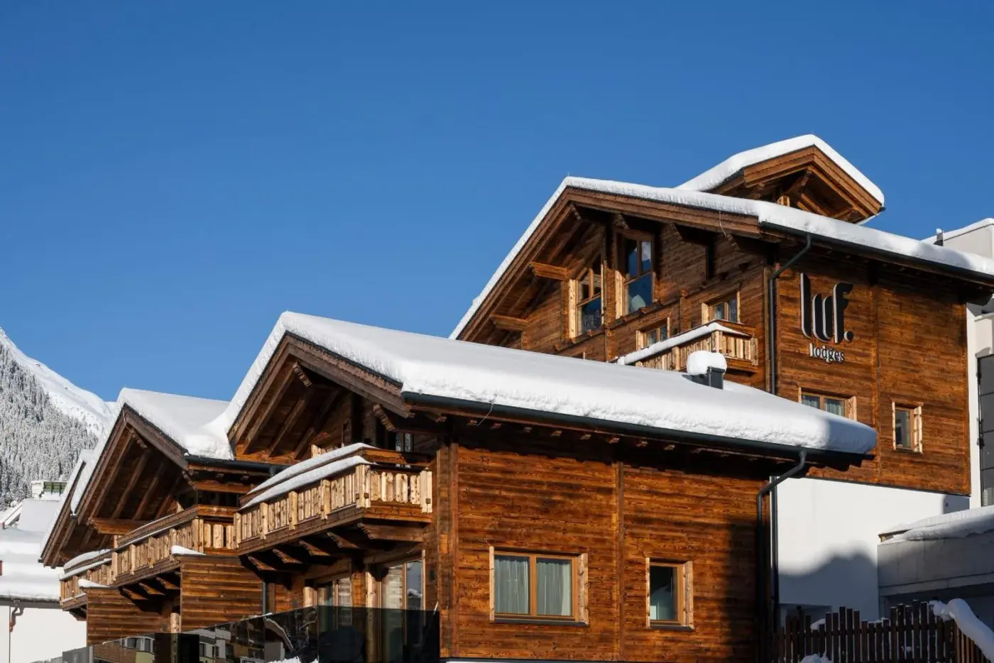 Timber lodges beneath a blue sky with snow on the roofs at Luf Lodges in Ischgl, Austria.