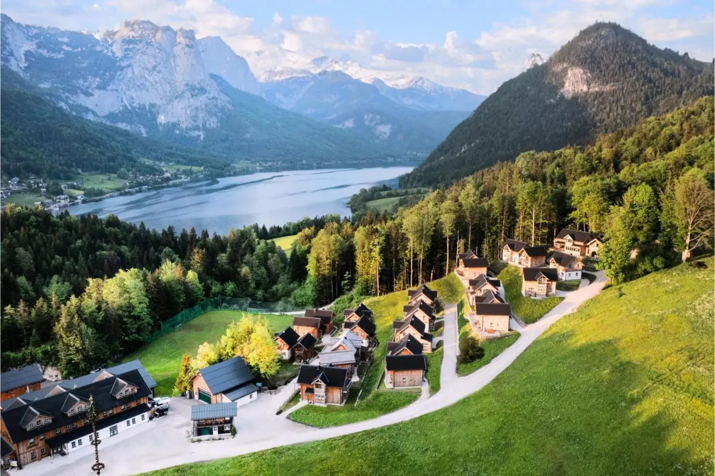 Aerial view of the chalet village beside a forest with lake and mountain views at Narzissendorf Zloam in Grundlsee, Austria.