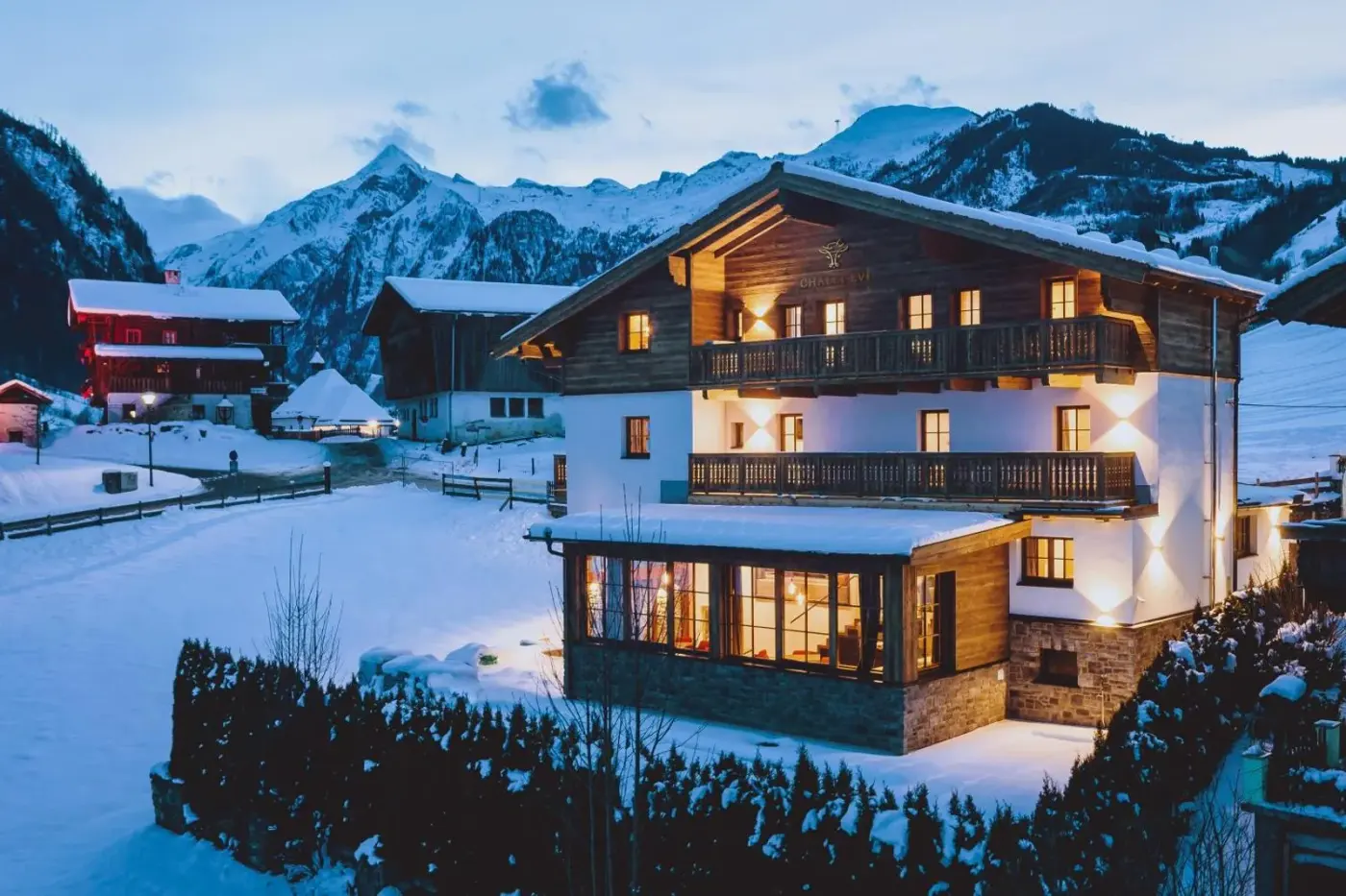 Illuminated chalet with timber balconies beneath snowy mountains at dusk at Chalet Evi in Kaprun, Austria.
