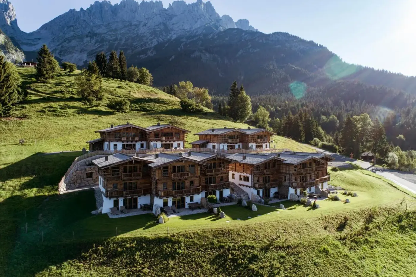 Timber chalet village on a green hillside beneath rocky mountain peaks at Wochenbrunner Chalets in Ellmau, Austria.